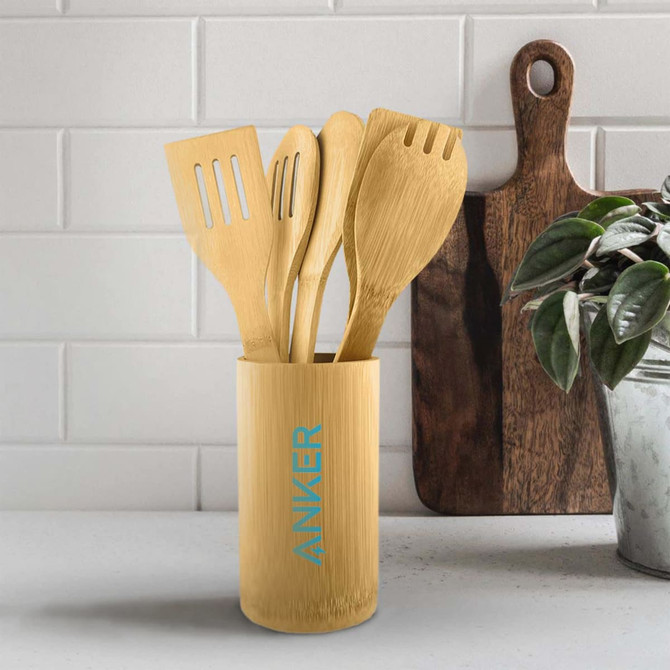 A bamboo cutlery set consisting of various utensils, displayed in a cylindrical holder, on a countertop with a plant and cutting board.