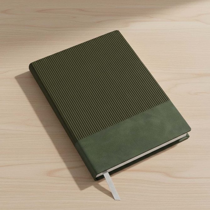 A black A5 notebook with a textured cover, resting on a wooden desk beside a window.