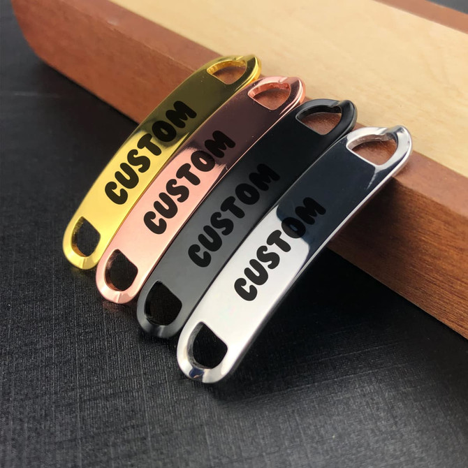 Four Smart Shoelace Charms in gold, rose gold, black, and silver, featuring a logo, arranged on a wooden surface.