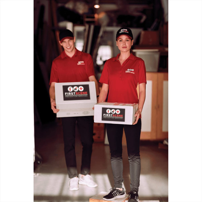 A pair of individuals wearing red polo shirts and caps, holding boxes in a warehouse setting.