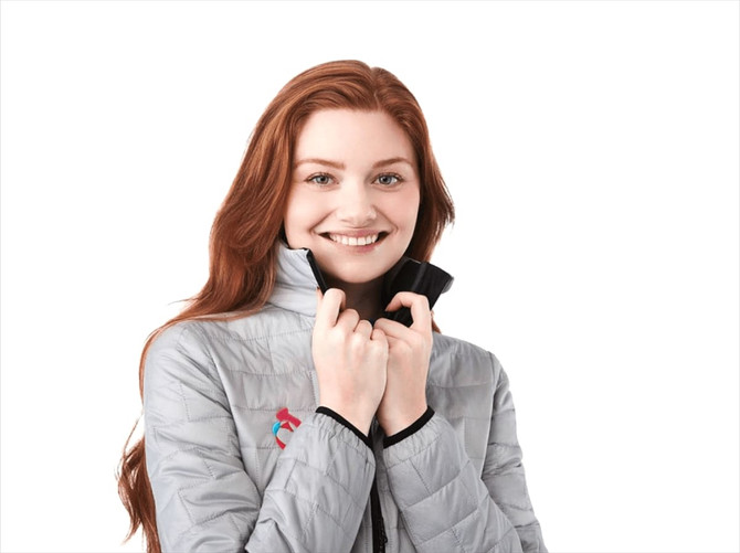 A woman wearing a grey lightweight insulated puffer jacket with a logo, smiling and adjusting the collar.