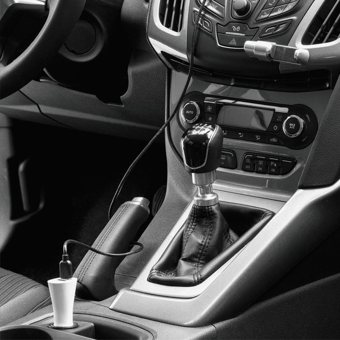 A dual car charger in white, plugged into a vehicle's centre console, next to the gear shift and dashboard controls.