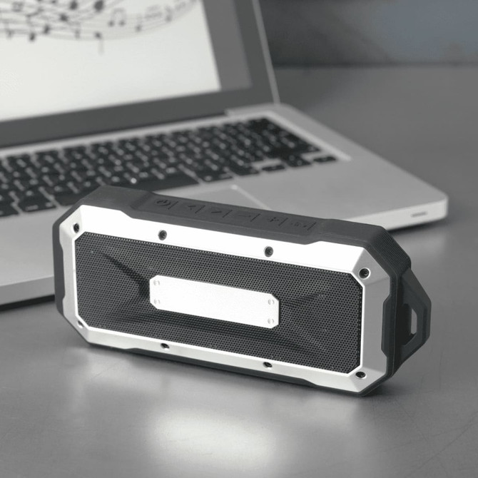 A rugged black and silver waterproof Bluetooth speaker placed beside a laptop on a metallic surface.