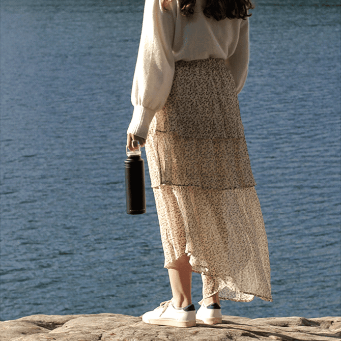 A drink bottle with a black exterior, held by a person in casual attire, near a body of water.
