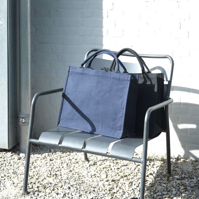 Navy blue recycled tote bags are displayed on a metal chair against a light brick wall. One bag is slightly behind the other.