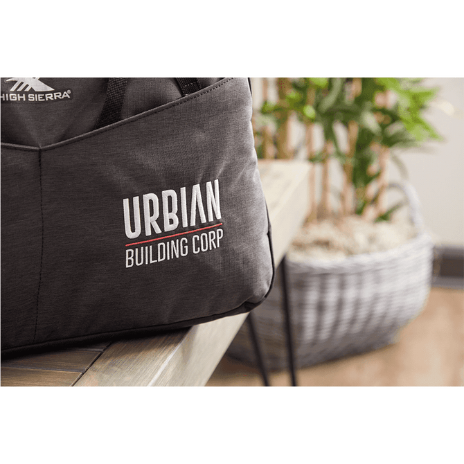 A dark grey duffel bag with a logo, placed on a table beside a potted plant and woven basket.