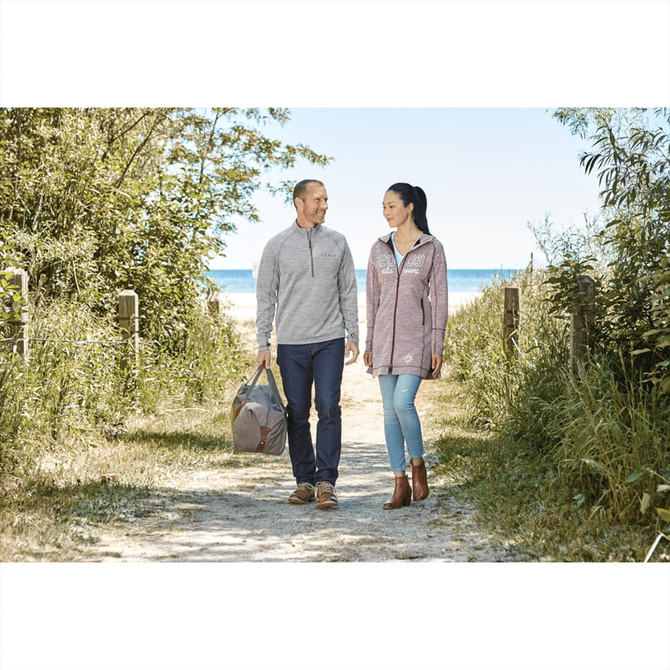 A man and woman walk along a sandy path wearing grey and maroon knitted jumpers, with a bag in hand and trees nearby.