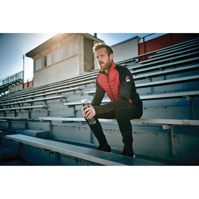 A man sits on stadium bleachers wearing a red and black insulated jacket, holding a drink bottle.
