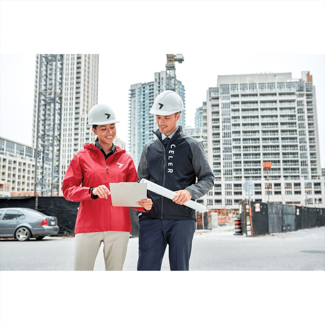 A woman in a red rain jacket and a man in a black jacket stand outside at a construction site, both wearing hard hats.