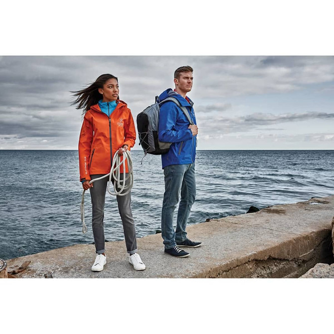 A woman in an orange softshell jacket and a man in a blue softshell jacket stand by the water, featuring a backpack.