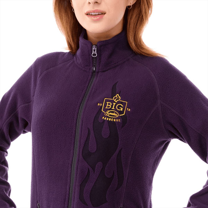 A women's polyfleece jacket in purple featuring a logo and black flame design on the front.