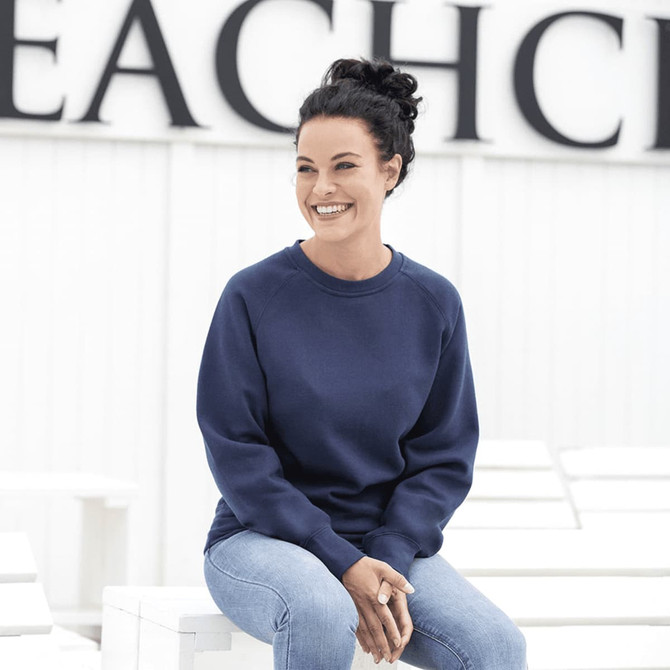 A woman sits wearing a navy blue fleece crew jumper, paired with light blue jeans, smiling against a neutral background.