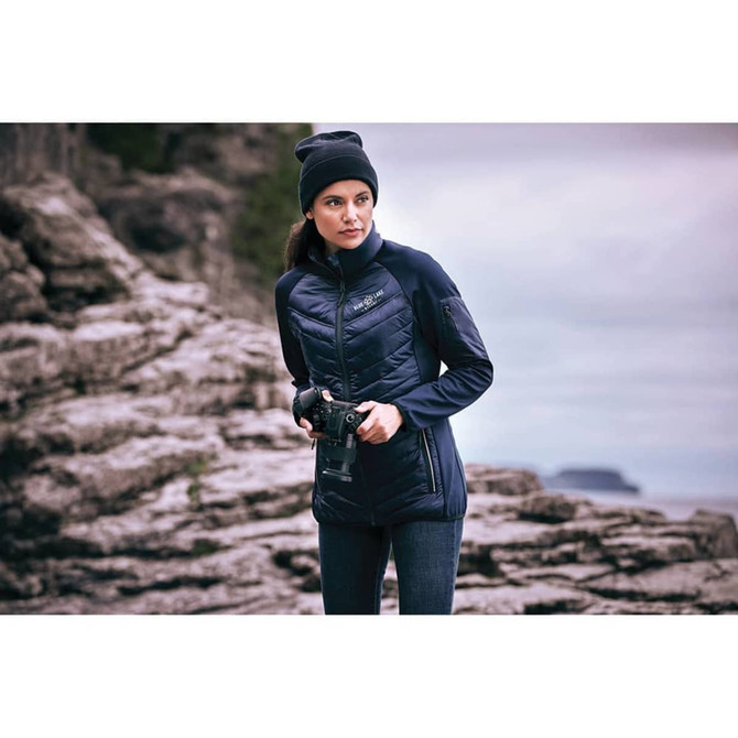 A woman wearing a navy insulated jacket, standing on rocky terrain, holding a camera. The jacket features a logo.