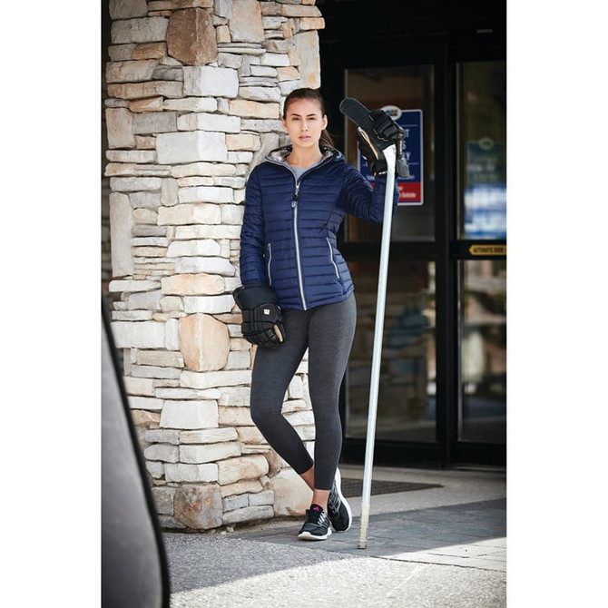A woman wearing a navy packable insulated jacket, black leggings, and sports gloves, standing by a stone wall.