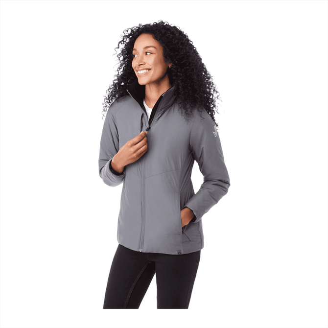 A woman is wearing a grey insulated puffer jacket with a zippered front and pockets, featuring a logo.