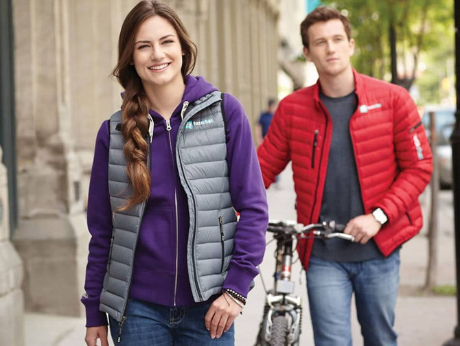 A woman in a grey puffy vest with a purple hoodie stands outdoors, smiling, with a man in a red puffy jacket nearby.