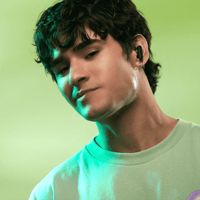 A person with dark hair is wearing black earbuds against a green background. The image features a logo on the earbuds.