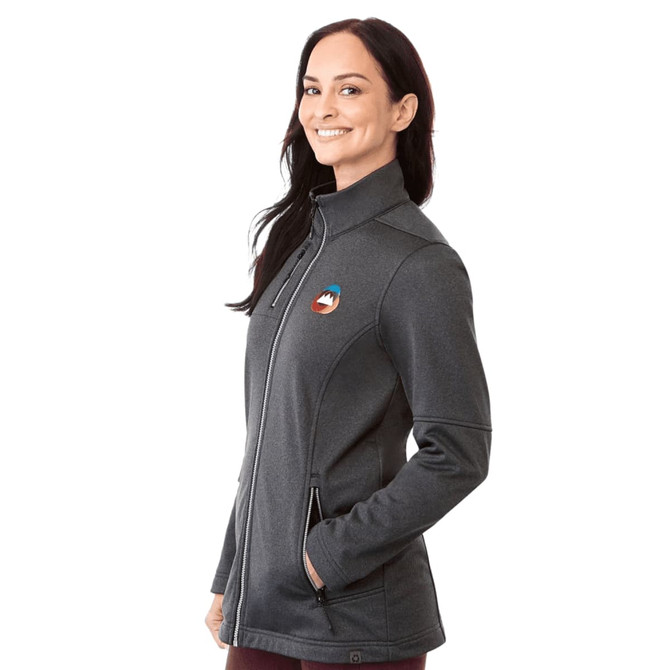 A woman wearing a grey softshell jacket with a logo, featuring a zip-up front and side pockets.