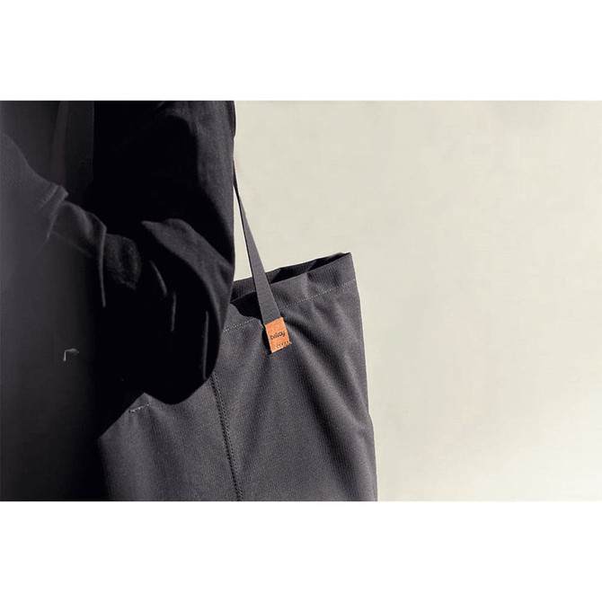A dark grey tote bag with a branding tag, held by a person in black clothing against a neutral background.