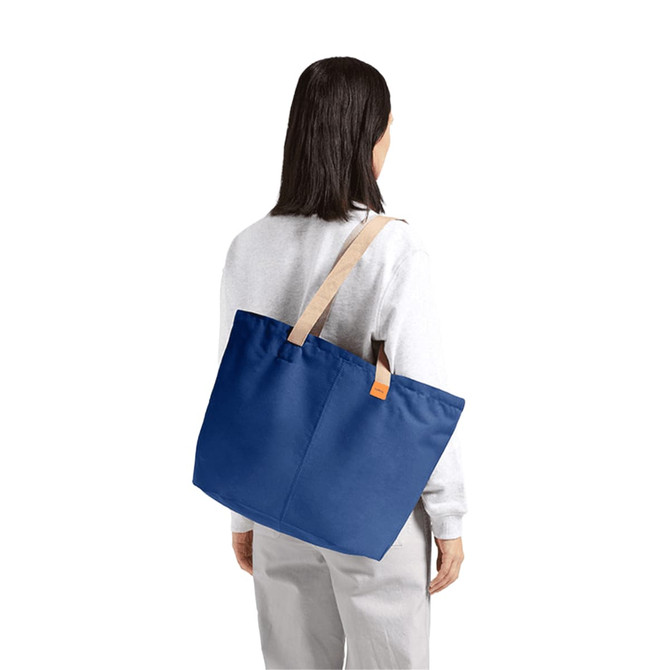 A blue tote bag with a light tan strap, being held by a person wearing a light grey sweater.