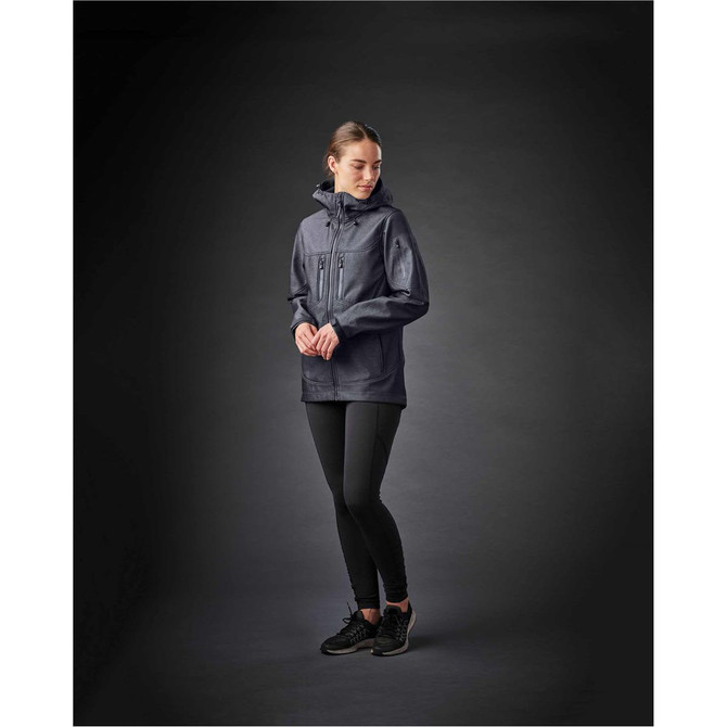 A woman wearing a dark grey softshell jacket, paired with black leggings and black shoes, on a dark background.