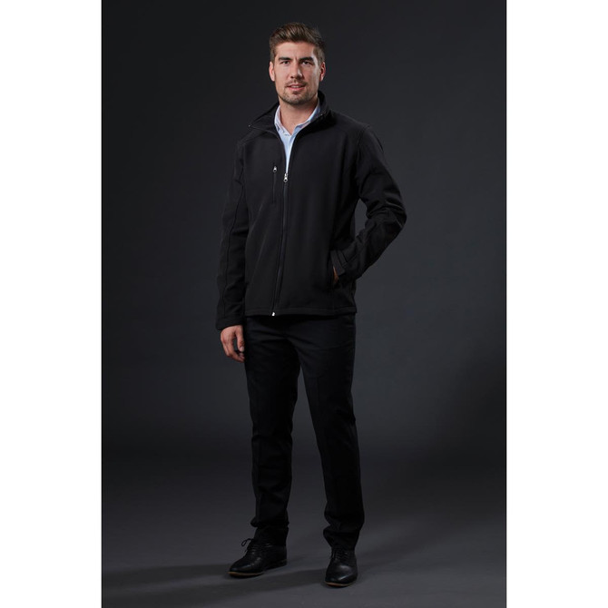 A man stands in a black softshell jacket, paired with black pants and shoes, against a dark background.