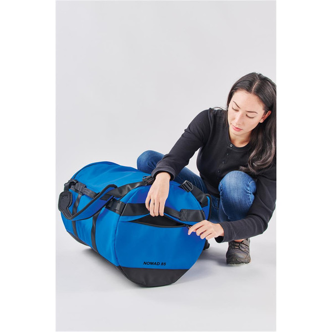 A blue duffle bag is being unzipped by a person, featuring black accents and a branding logo.