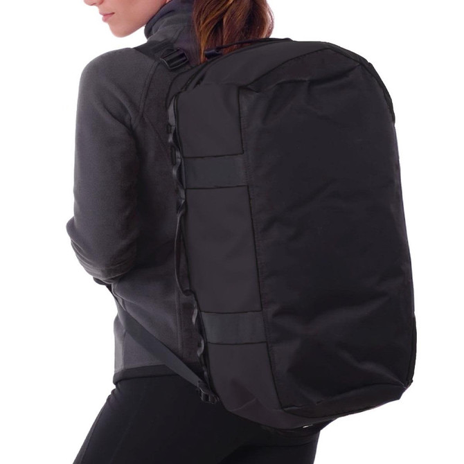 A black duffle bag is being worn on the back of a person in a grey jacket. The bag features a logo.