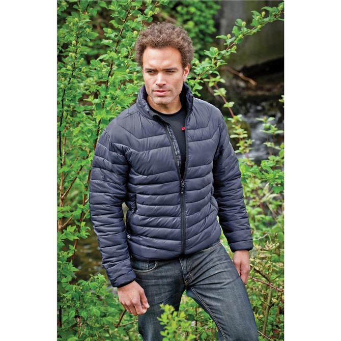 A man wearing a dark navy puffer jacket stands amidst lush greenery, with a creek visible in the background.