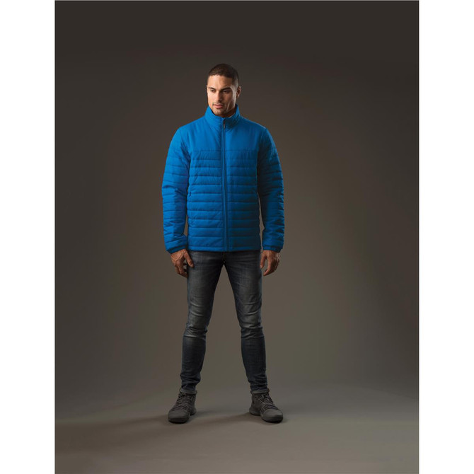 A man stands wearing a blue quilted jacket, paired with jeans and boots, with a neutral background.