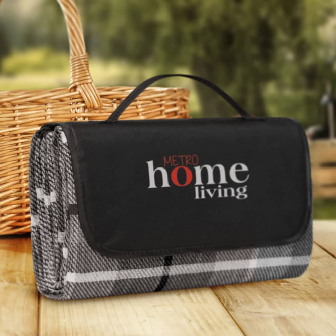 A folded picnic blanket in grey and black, featuring a handle and a logo on the front. A wicker basket is nearby.