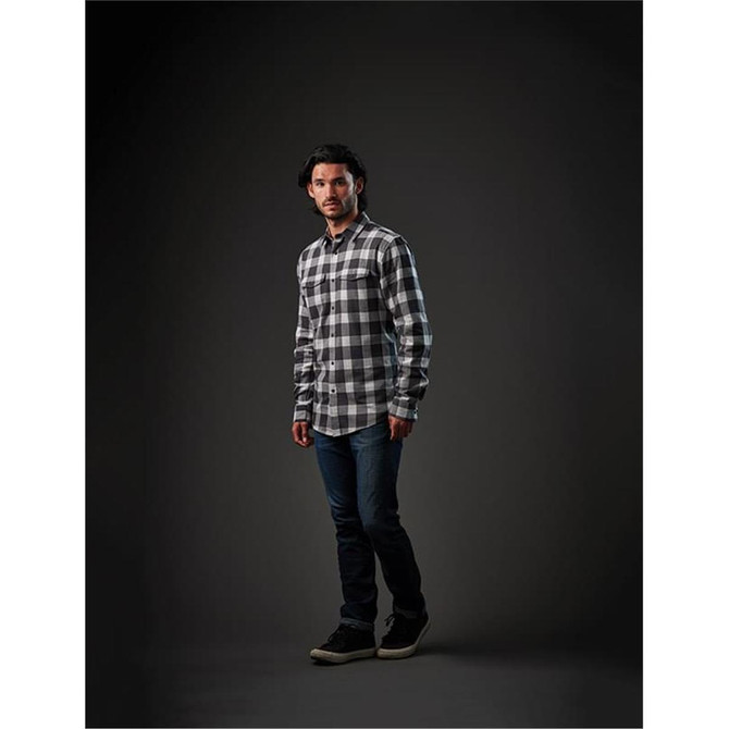 A man stands in a dark setting wearing a black and white checked long-sleeve button-up shirt and jeans.
