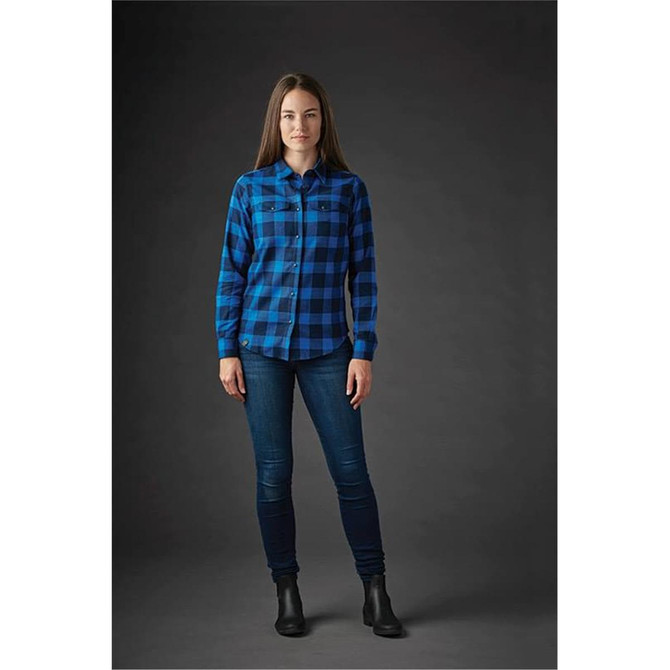 A woman stands wearing a blue and black checked button-up shirt and jeans, with a plain dark background.