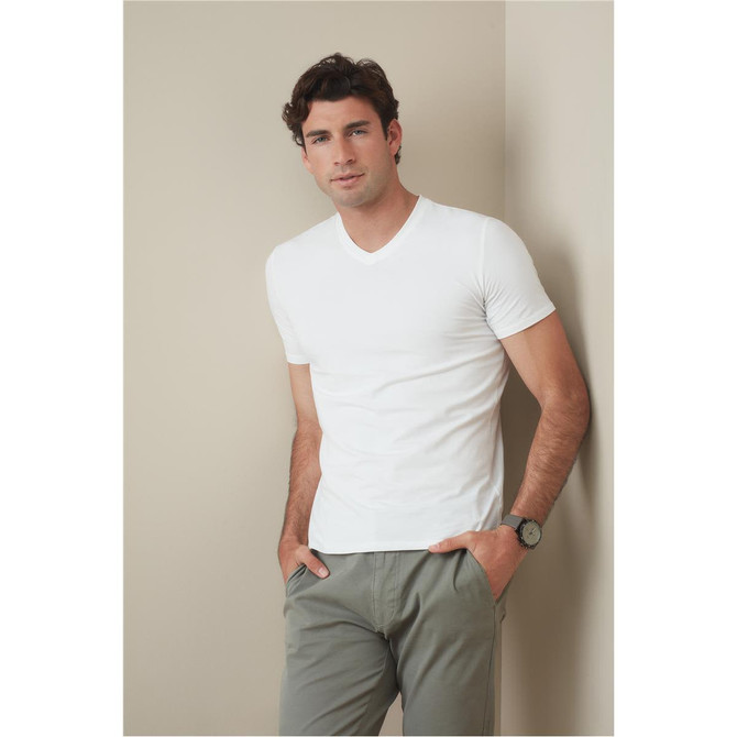 A man in a white V-neck t-shirt stands against a neutral background, hands in pockets, wearing casual pants.