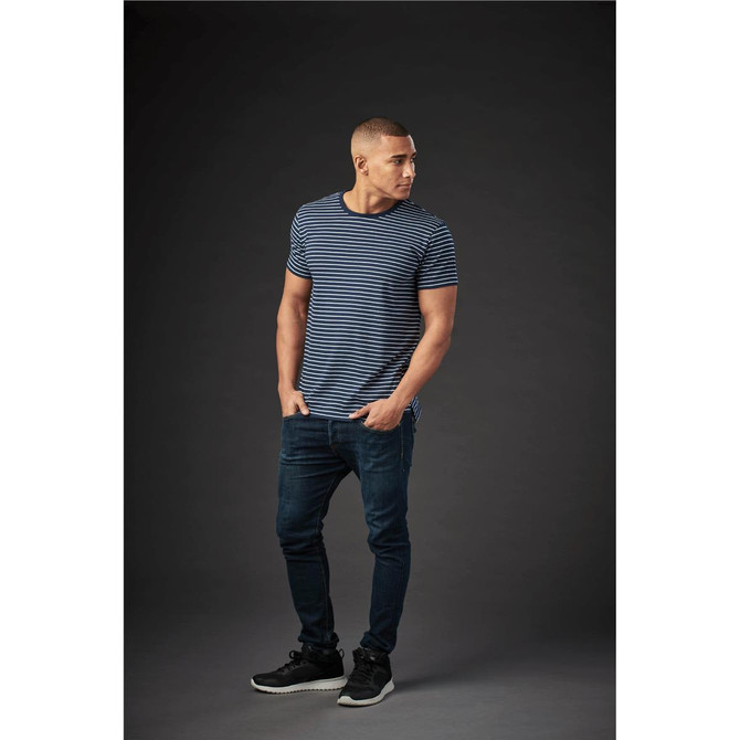 A man stands casually wearing a blue and white striped crew neck t-shirt and dark jeans, with a logo on the shirt.