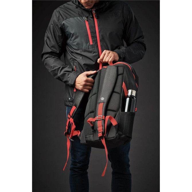 A black and red backpack is held by a person wearing a black jacket, featuring a side pocket for a drink bottle.