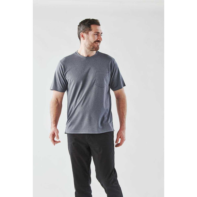 A man stands wearing a grey short sleeve t-shirt with a pocket, paired with dark pants. The shirt features a logo.