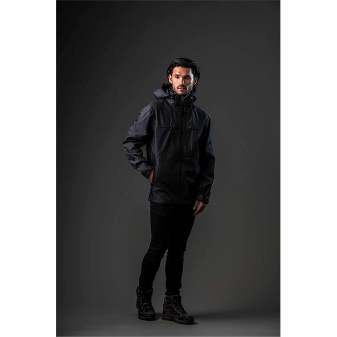 A man wearing a black softshell jacket stands against a dark background. The jacket features a hood and has a logo.