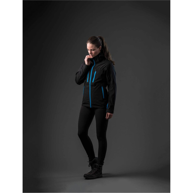 A woman wearing a black jacket with blue accents, styled with black leggings and boots, stands against a dark background.