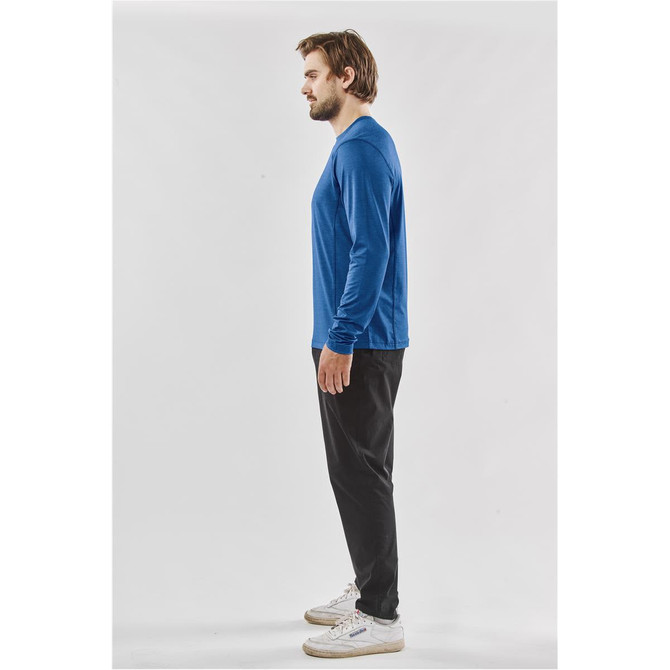 A man stands sideways wearing a blue long-sleeve crew neck shirt and black pants, with a logo visible.
