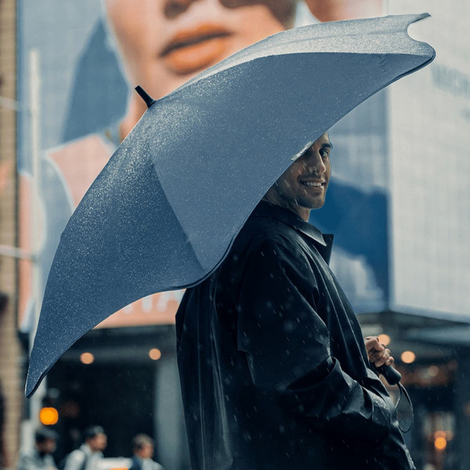A navy BLUNT Exec Umbrella held by a person in a raincoat, with city buildings in the background.