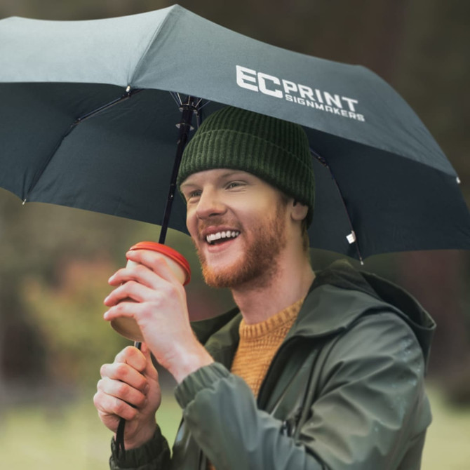 A man holding a red coffee cup under a dark green tri-fold umbrella, wearing a beanie and a jacket.