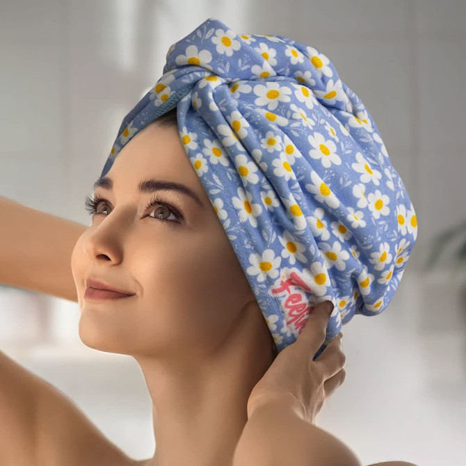 A woman wearing a light blue hair towel decorated with white daisies and yellow accents, featuring a logo.