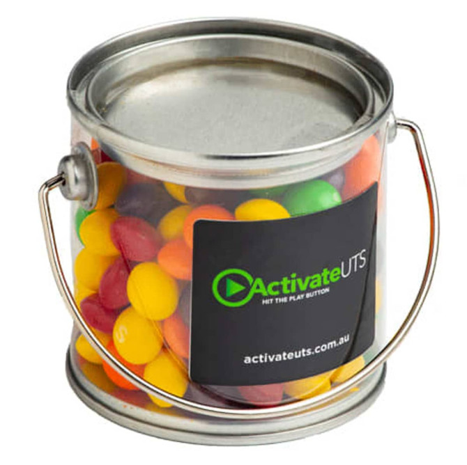 A small clear PVC bucket filled with vibrant Skittles in various colours, featuring a metal handle and a logo.