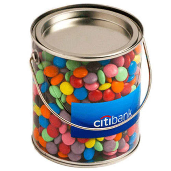 A clear PVC bucket filled with colourful choc beans, featuring a metal lid and a handle. Includes branding on the front.
