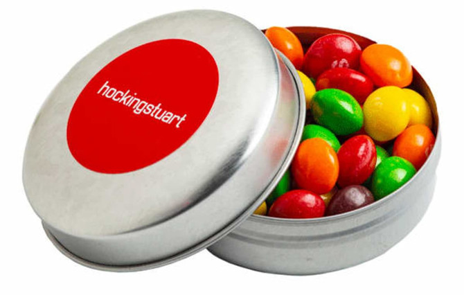 A silver tin filled with colourful Skittles—red, orange, yellow, green, and purple. It features a round logo on the lid.