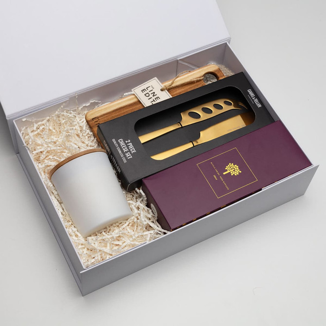 A gift box containing a cheese board, gold cheese knives, and a ceramic container, all set in white paper fill.