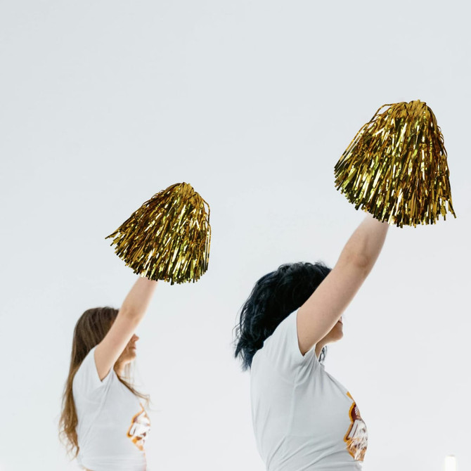 Gold cheerleading pom poms held up by two individuals wearing white t-shirts, with a simple background.