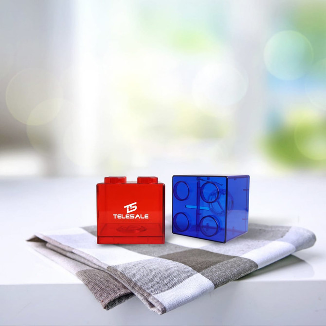 A red and a blue square block coin bank placed on a grey and white checked cloth. The blue block has a logo.