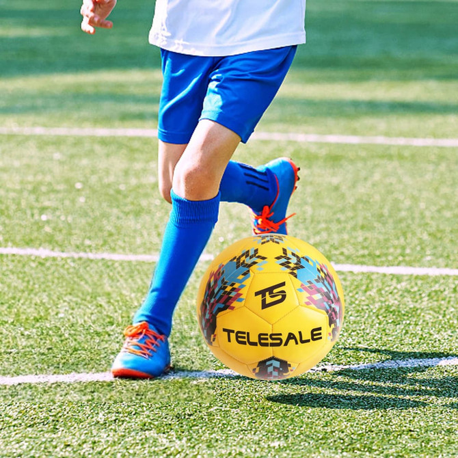 A training soccer ball in yellow with a colourful design, placed on green grass, as a child runs by.